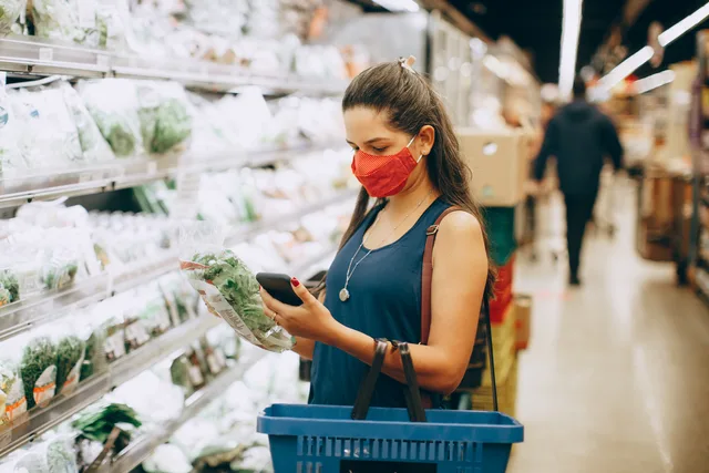 A woman wearing a mask shops for groceries in a supermarket, checking products with her phone. Regionale Preisunterschiede vergleichen: Datenquellen und methodische Grenzen
